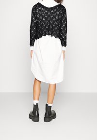 Schwarzer gestrickter, kurzer Pullover mit offenem Muster, getragen über einem weißen Hemdkleid; kombiniert mit schwarzen Stiefeletten und weißen Socken.
