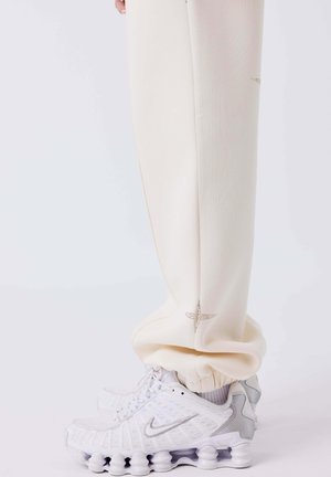 Onderbenen in crèmekleurige broeken met geborduurde sterren, gecombineerd met witte en grijze Nike-sneakers met een uniek zoolontwerp.