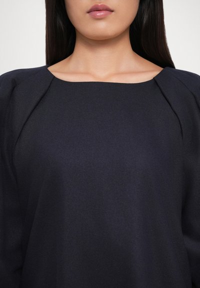 Woman with straight dark hair wearing a navy blue square-neck blouse with gathered shoulder details, shown from neck to chest.