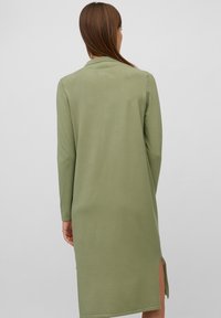 Femme aux cheveux longs et bruns portant une robe olive verte à manches longues et longueur genou avec une fente latérale, vue de dos sur un fond uni.