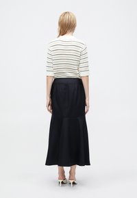Blonde woman wearing a white striped short-sleeve top, black long flared skirt, and white heeled shoes, standing facing away on white background.