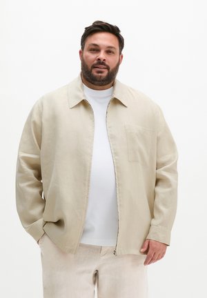 Brodaty mężczyzna w beżowej kurtce na zamek błyskawiczny, białej koszuli i pasujących beżowych spodniach, stojący z jedną ręką w kieszeni na białym tle.