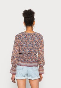 Blusa floral com mangas longas e bufantes, cintura franzida e um padrão multicolorido em azul, laranja e vermelho. Combinada com calções de ganga claros.