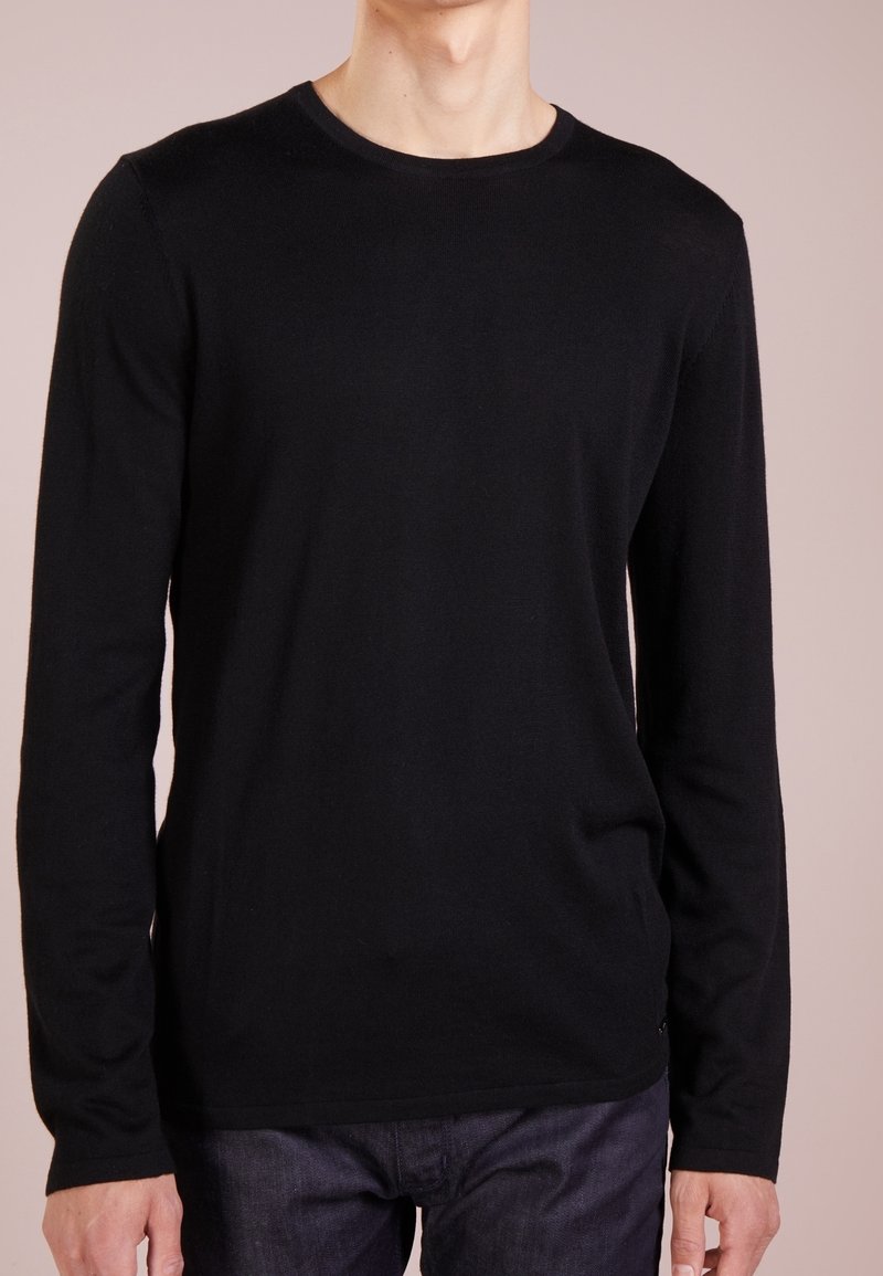 Uomo che indossa una maglietta nera a maniche lunghe con collo rotondo e jeans blu scuro, su uno sfondo beige semplice.