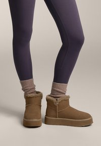 Brązowe zamszowe botki za kostkę z futrzanym obszyciem, gumową podeszwą i gładkim wykończeniem, połączone z lawendowymi, prążkowanymi skarpetkami i ciemnymi legginsami.