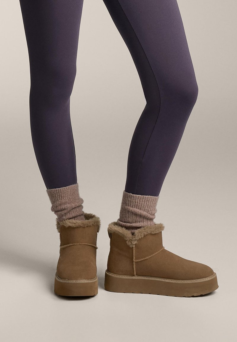 Brązowe zamszowe botki za kostkę z futrzanym obszyciem, gumową podeszwą i gładkim wykończeniem, połączone z lawendowymi, prążkowanymi skarpetkami i ciemnymi legginsami.