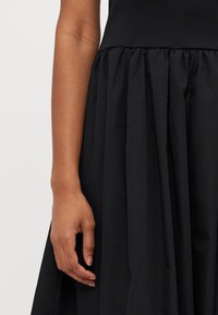 Svart kjole med tettsittende bol og en plissert, flytende skjørt. Stoffet virker glatt med en matt overflate. Hånden hviler på skjørtet.