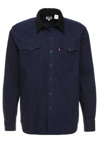 Tmavě modr�á džínová košile s černým manžestrovým límečkem, dvěma náprsními kapsami, zapínáním na knoflíky vpředu a dlouhými rukávy. Texturovaná tkanina s pevnými švy.