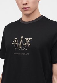 Νέος άνδρας που φοράει μαύρο t-shirt Armani Exchange με κεντημένο λογότυπο A|X με πολλαπλά στιλ στο στήθος.
