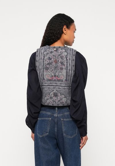 Γκρι καπνιστό κοντό γιλέκο με περίπλοκες λουλουδάτες λεπτομέρειες, συνδυασμένο με ένα σκοτεινό μακρύ μανίκι και ψηλόμεσα μπλε τζιν παντελόνια.