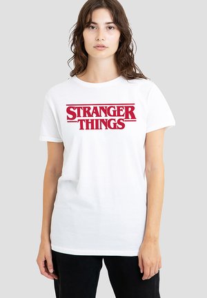 Young woman with wavy brown hair wearing a white t-shirt with red "Stranger Things" text, standing against a plain light gray background.