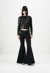 Femme portant un pull en tricot noir, un jean évasé noir, des lunettes de soleil noires et des sandales à bout ouvert, se tenant contre un fond blanc.