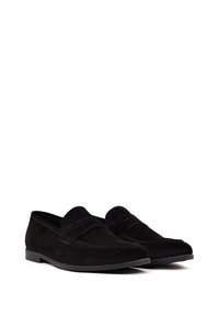 Mocassins en suède noir avec une texture lisse, présentant des coutures décoratives et une bande surélevée sur le dessus. Semelle plate avec des détails subtils.