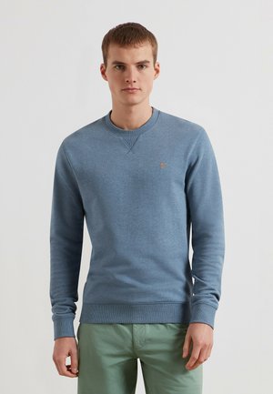 Mężczyzna ubrany w niebieską bluzę z długim rękawem z małym pomarańczowym logo na piersi i jasnozielone spodnie na jednolitym białym tle.