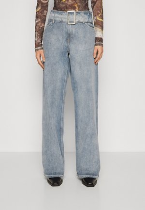 Jean bleu clair à jambes larges avec taille haute, grande ceinture avec boucle carrée, porté avec des chaussures noires pointues et un haut à manches longues à motifs.