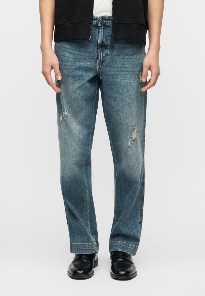 Άτομο που φοράει ξεθωριασμένα μπλε τζιν με σκισμένες περιοχές και μαύρα δερμάτινα παπούτσια, που στέκεται μπροστά σε έναν απλό φωτεινό φόντο.