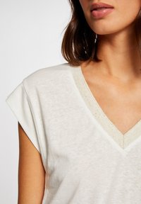 Witte v-hals t-shirt gemaakt van een textuurstof, met korte mouwen en een metallic gouden rand langs de halslijn.