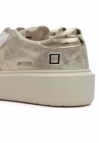 Les baskets présentent une tige en or métallique clair avec une finition texturée, des lacets blancs et une semelle épaisse blanche. Un petit logo est visible sur le côté.
