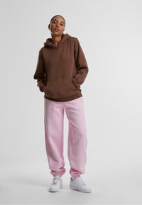 Hnedá mikina s kapucňou a predným vreckom, skombinovaná s svetloružovými teplákmi. Biele tenisky dopĺňajú outfit.