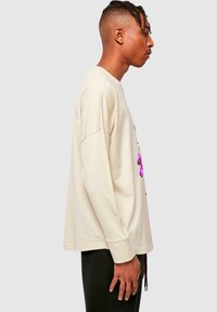 Beige Baumwoll-T-Shirt mit langen Ärmeln, vorderem Grafikdruck und Rundhalsausschnitt. Abgerundeter Saum, lockere Passform und tief sitzende Schulternähte.