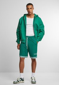Zöld zipzáros kapucnis felső, fehér ujjatlan póló, és zöld rövidnadrág fehér részletekkel. Sportos design, laza szabás és húzózsinórral ellátott dér.