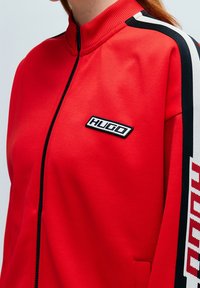 Červená zipová bunda s vysokým límcem, s černobílými pruhovanými detaily a logem HUGO na hrudi. Hladká textilie.