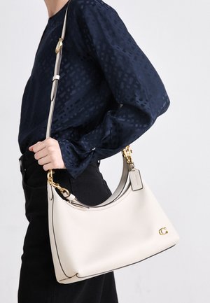 Een beige leren handtas met een donkere afwerking, een gebogen silhouette, gouden accenten en een afneembare schouderriem, vastgehouden aan de zijkant tegen een donkere outfit.