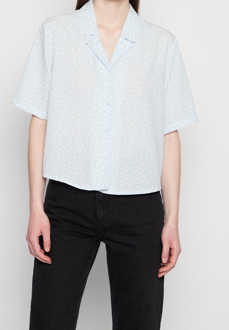 Short-sleeve, light blue blouse with a floral pattern, button front, and a relaxed fit. Made of soft fabric with a collar and a cropped design.