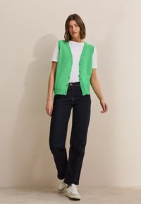 Grön stickad väst med knappar framtill, bärs över en vit t-shirt. Kombinerad med mörka jeans och vita sneakers. Slät textur och åtsittande design.