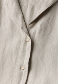 Beige linneskjorta med en mjuk textur, försedd med krage, en rad små knappar och subtila sömmar.
