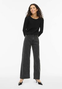 Suéter negro con cuello redondo, combinado con jeans de pierna ancha y talle alto en color antracita. Modelo lleva tacones negros puntiagudos, posando sobre un fondo liso.