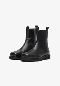 Botas de tobillo de cuero negro con un acabado suave, cremallera lateral y tiradores. Suela de goma resistente, detalles cosidos y un diseño clásico.
