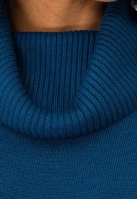 Gros plan d’une personne portant un pull à col roulé en maille côtelée bleu, montrant le cou et la partie supérieure de la poitrine.