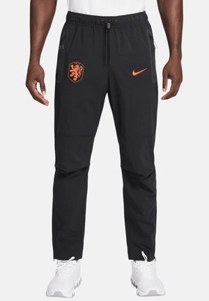 Czarne spodnie sportowe z pomarańczowym herbem lwa i logo Nike, kieszenie na zamek, elastyczna talia, noszone z białymi sneakersami przez stojącą osobę.