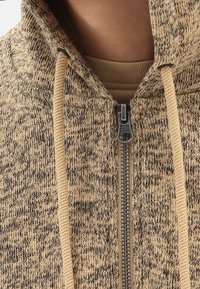 Beige a tmavě hnědá mikina s kapucí s texturovaným pleteným vzorem, obsahující zip vpředu a béžové šňůrky. Obsahuje kovový zip.