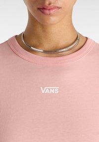 Růžové bavlněné tričko s kulatým výstřihem a bílým logem "VANS". Kovový choker náhrdelník ze stříbra se nachází kolem krku. Hladký povrch.