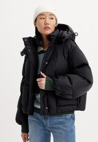 Black puffer jacket with a hood, featuring a quilted design, snap buttons, and ribbed green cuffs, worn over a light blue sweater.