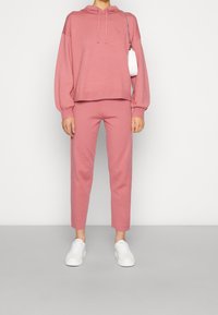 Felpa con cappuccio rosa e pantaloni tapered abbinati, realizzati in materiale morbido. Abbinata a sneakers bianche, l'outfit presenta un design minimalista.