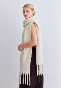 Wollen sjaal in crèmekleur, met franjeranden en een logolabel. Zachte textuur, lang model dat elegant om de schouders is gedrapeerd.