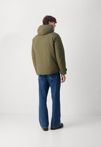 Olivengrøn polstret jakke med hætte, med en glat overflade, afslappet pasform og manchet detaljer, parret med blå flared jeans.