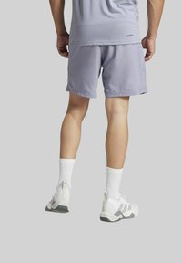 Man in een lichtgrijze sportshort, witte sokken en witte Adidas-sneakers, staand voor een effen grijze achtergrond.