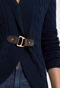 Tamsiai mėlynas megztas kardiganas su ribboto audinio pelerinos apykakle ir tamsiai rudos odos sagtimi, derinamas su mėlynomis džinsinėmis kelnėmis.