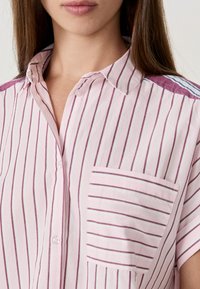 Femme portant une chemise boutonnée à rayures verticales rose et blanche avec une poche poitrine, un col et des accents maroon aux épaules.