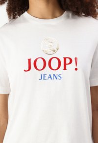 Balta medvilninė marškinėlių su trumpomis rankovėmis, ant krūtinės užrašas "JOOP!" raudona spalva ir "JEANS" mėlyna spalva, taip pat auksinė apskrita logotipo dizainas.