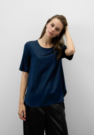 Donna con lunghi capelli castani che indossa orecchini di perle, maglietta blu navy a maniche corte e pantaloni neri, che si tocca i capelli davanti a uno sfondo di muro chiaro e uniforme.