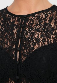 Μαύρο τοπ από δαντέλα με λουλουδάτα μοτίβα, στρογγυλή λαιμόκοψη και σειρά από πέντε στρογγυλά μαύρα κουμπιά στο μπροστινό μέρος. Μαλακό, υφασμάτινο με υφή.