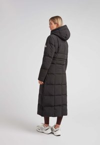 Abrigo negro largo tipo puffer con capucha, con un diseño acolchado y una cremallera en la parte inferior. Combinado con mallas y zapatillas deportivas.