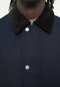 Veste bleu marine avec un col en velours côtelé noir, dotée de boutons-pression argentés et d'une texture de tissu résistante. Gros plan sur l'encolure.