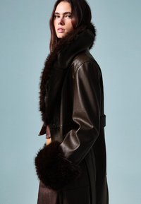 Hnědý kožený kabát s velkým kožešinovým límcem a manžetami. Má přiléhavý střih a zapínání na knoflík, s hladkou texturou.