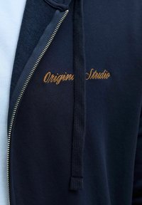 Marineblauwe zip-up hoodie met een gladde textuur. Bevat gouden geborduurde 'Original Studio' details, met contrasterende blauwe stof eronder.